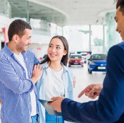 img-3-1 new A car dealership scene featuring a salesperson engaging with two customers while showcasing vehicles in the background.