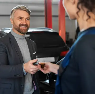 img-6-1-new A man in a suit hands car keys to a woman in a blue dress, surrounded by parked cars in a well-lit garage.