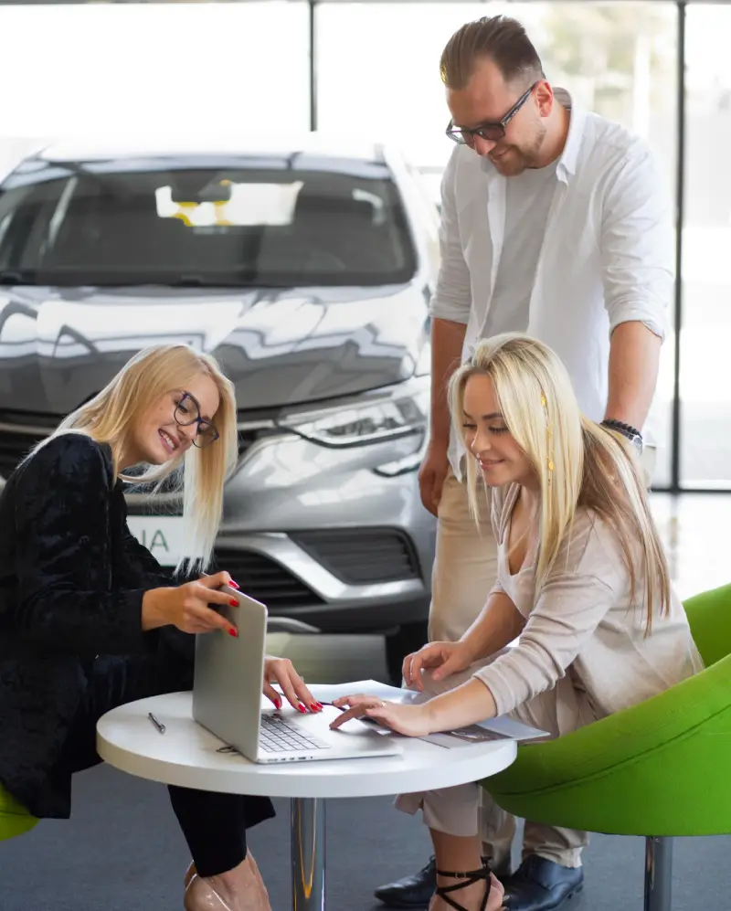Three individuals collaborate at a table with a laptop in a car dealership, while a sleek vehicle is prominently displayed in the background.